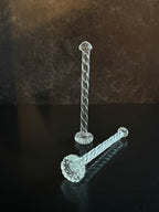Two clear twisted glass cocktail muddlers on a reflective black surface against a dark brown textured background.