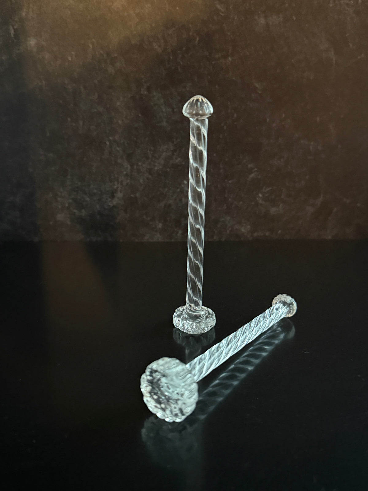 Two clear twisted glass cocktail muddlers on a reflective black surface against a dark brown textured background.