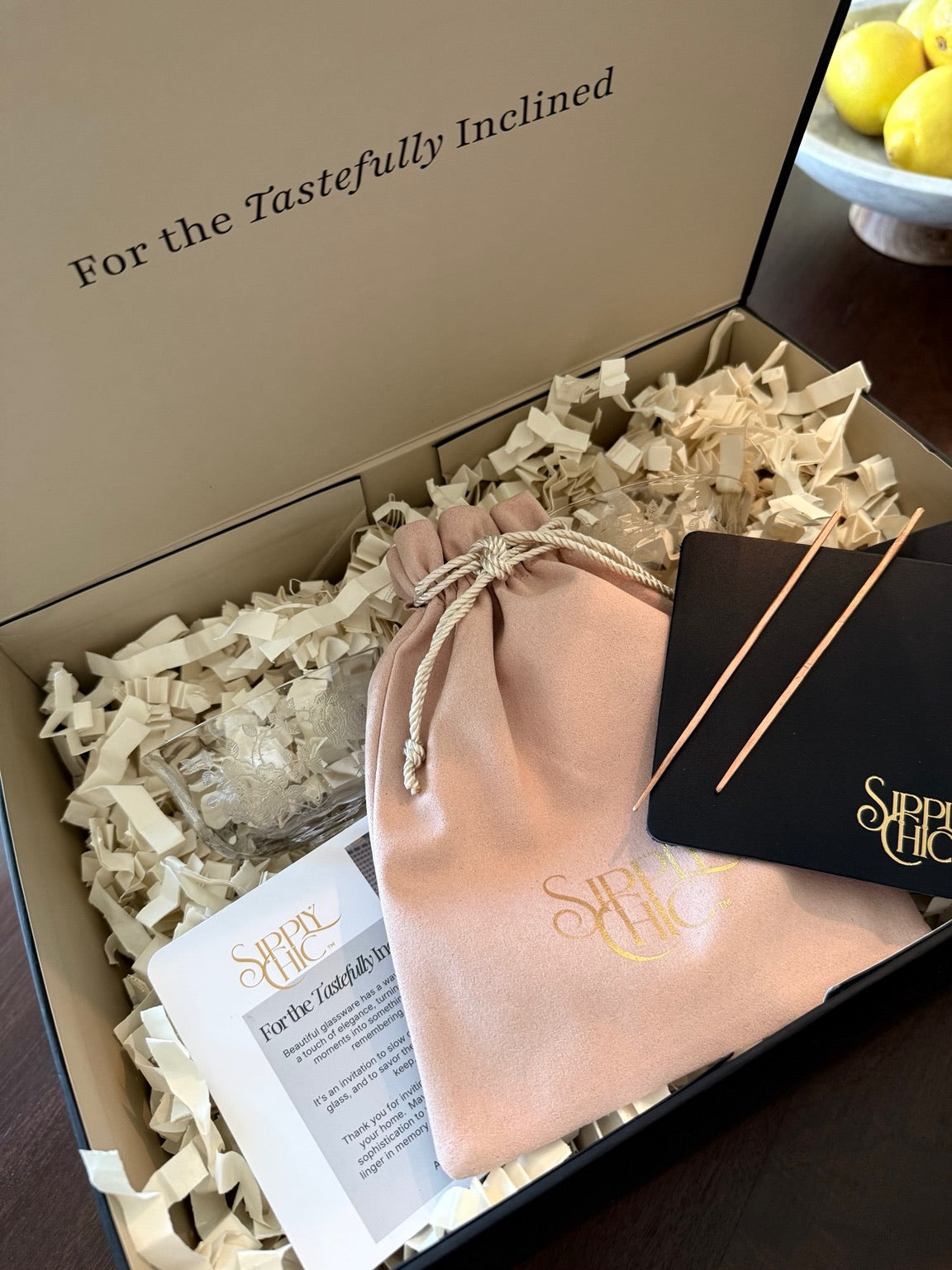 An open gift box containing a blush pink drawstring pouch with gold foil 'Sipply Chic' branding, pair of navy coasters, two copper cocktail picks, a printed card, and shredded paper filler. The inside lid of the box reads 'For the Tastefully Inclined'.