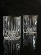 Two clear cut-crystal tumbler glasses with a vertical ribbed pattern and a faceted base, displayed on a reflective black surface against a dark textured background.