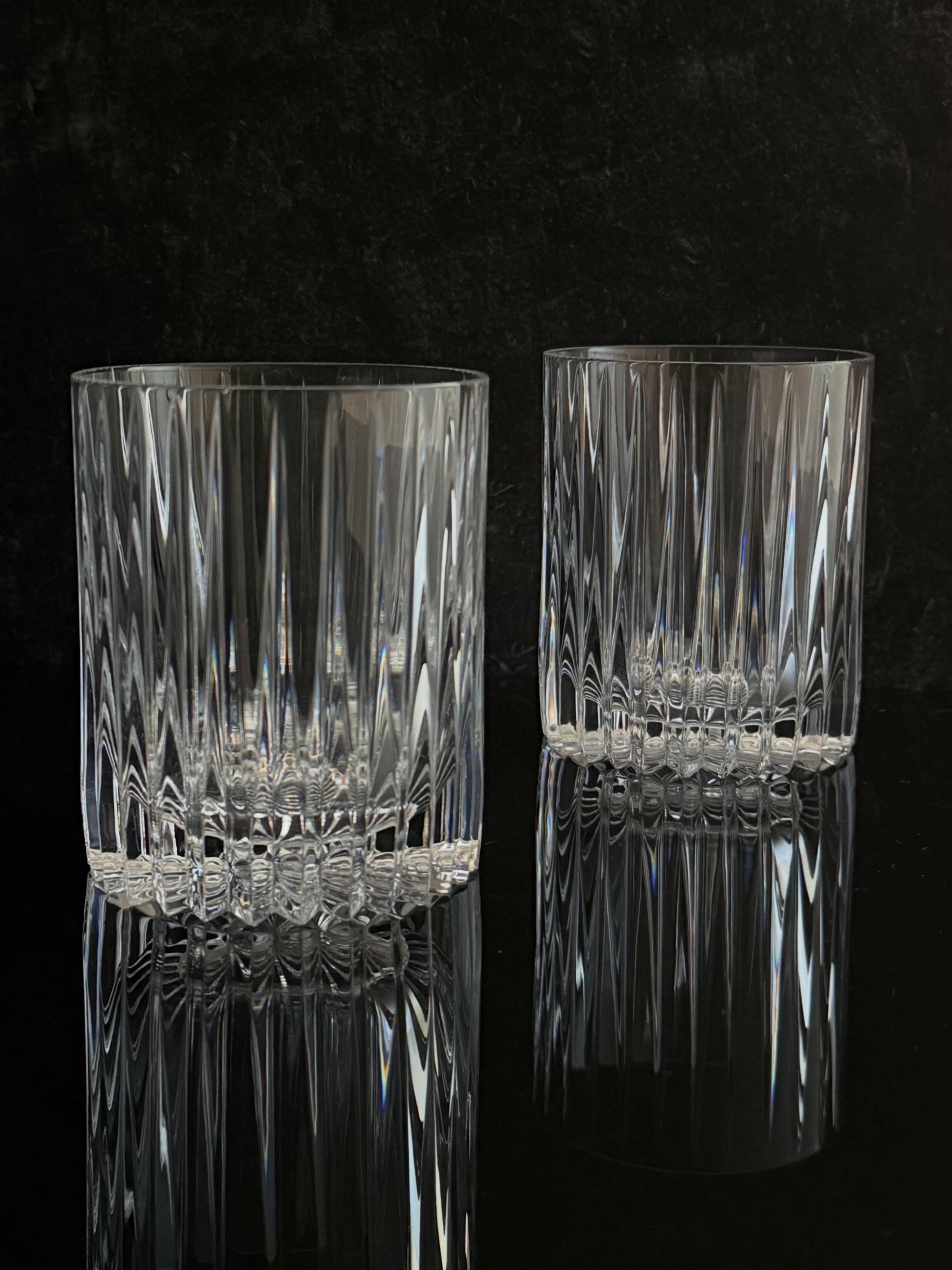 Two clear cut-crystal tumbler glasses with a vertical ribbed pattern and a faceted base, displayed on a reflective black surface against a dark textured background.