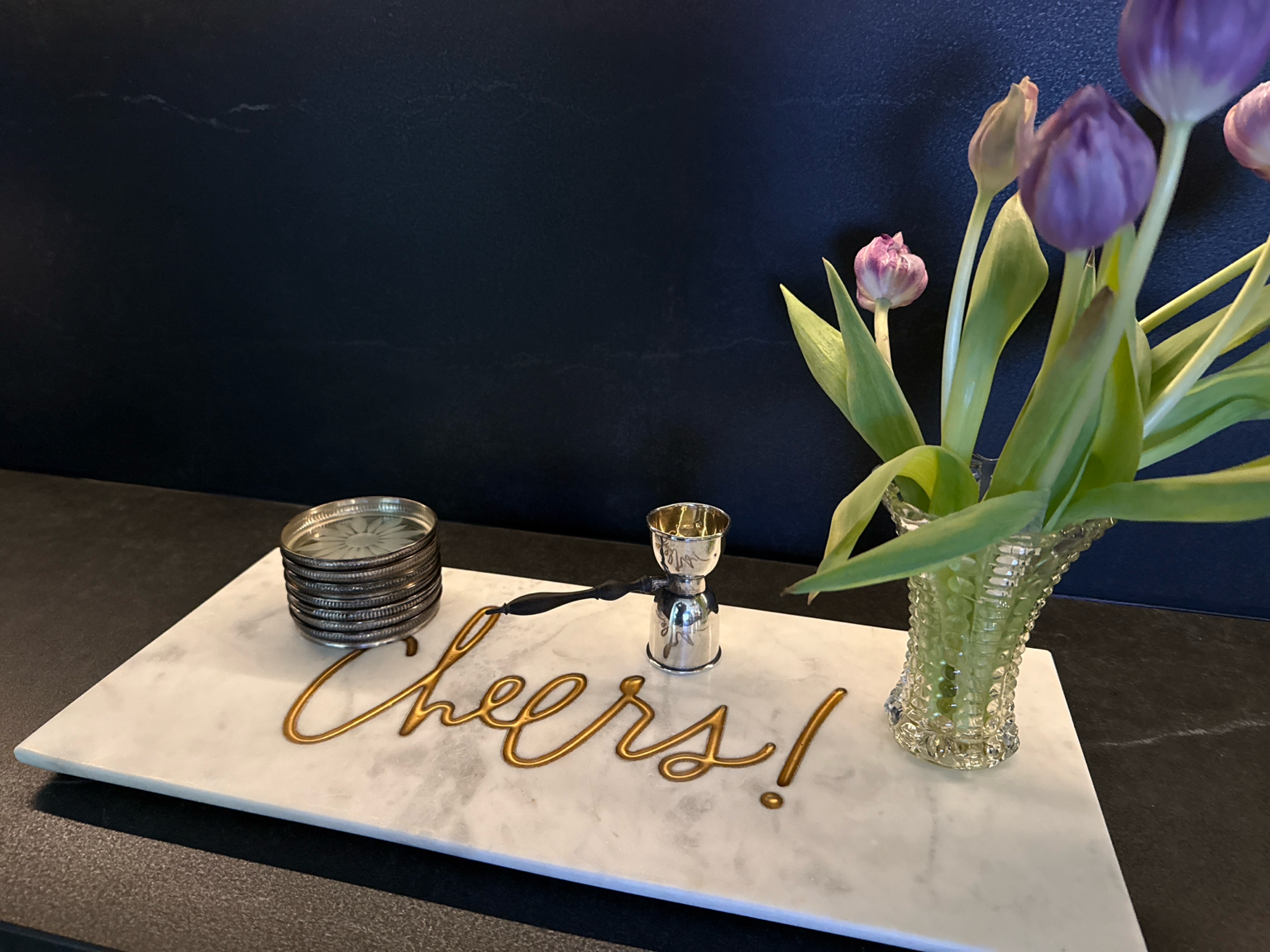 A white marble serving board on a dark table, featuring a gold wire script sign that says 'Cheers!', a stack of metal coasters, a small silver jigger, and a glass vase filled with purple tulips against a dark navy wall.