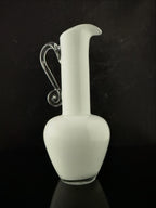 A white glass pitcher with a curved neck, a clear glass handle, and a small clear glass base, set against a dark black background.