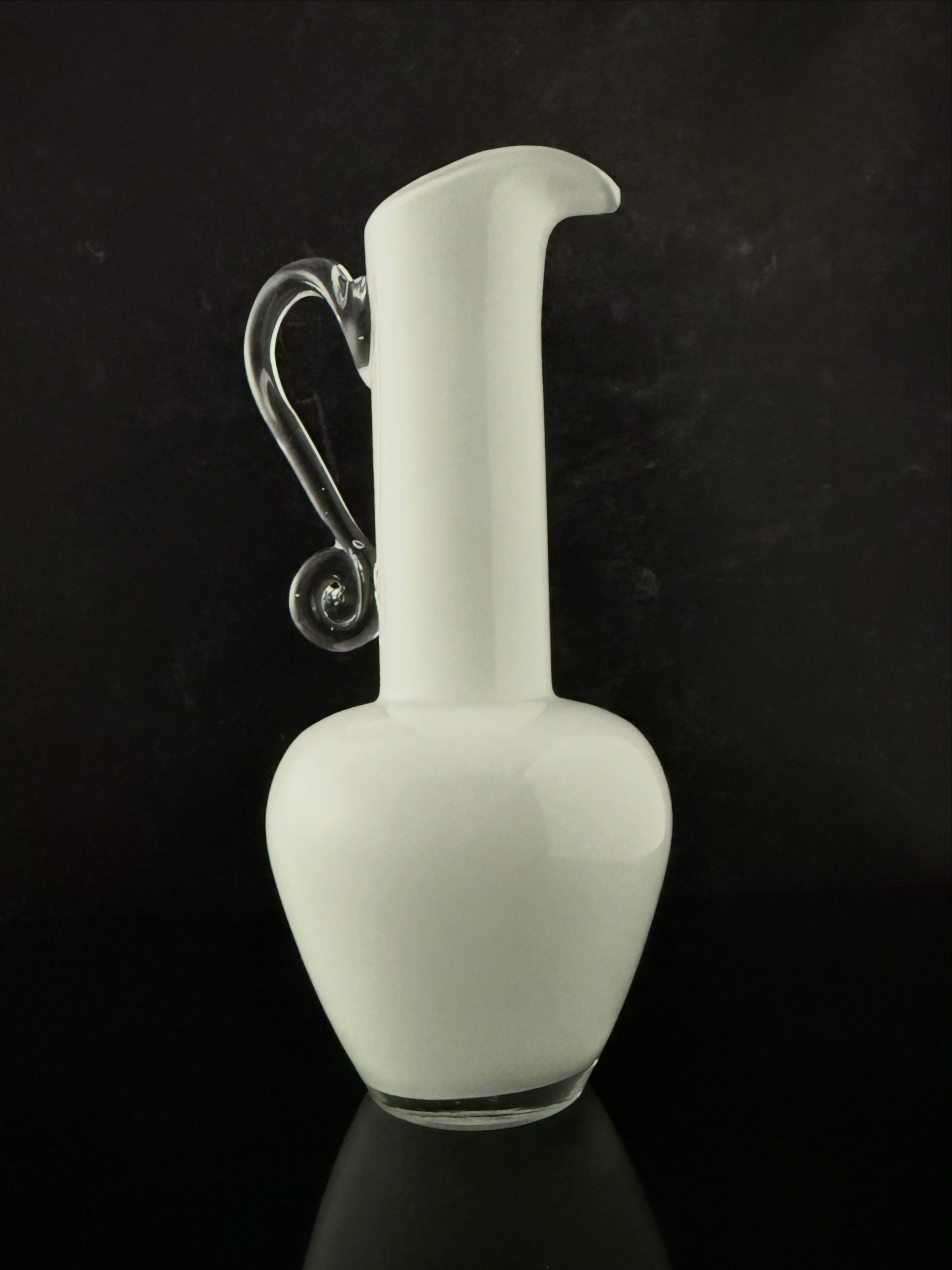 A white glass pitcher with a curved neck, a clear glass handle, and a small clear glass base, set against a dark black background.