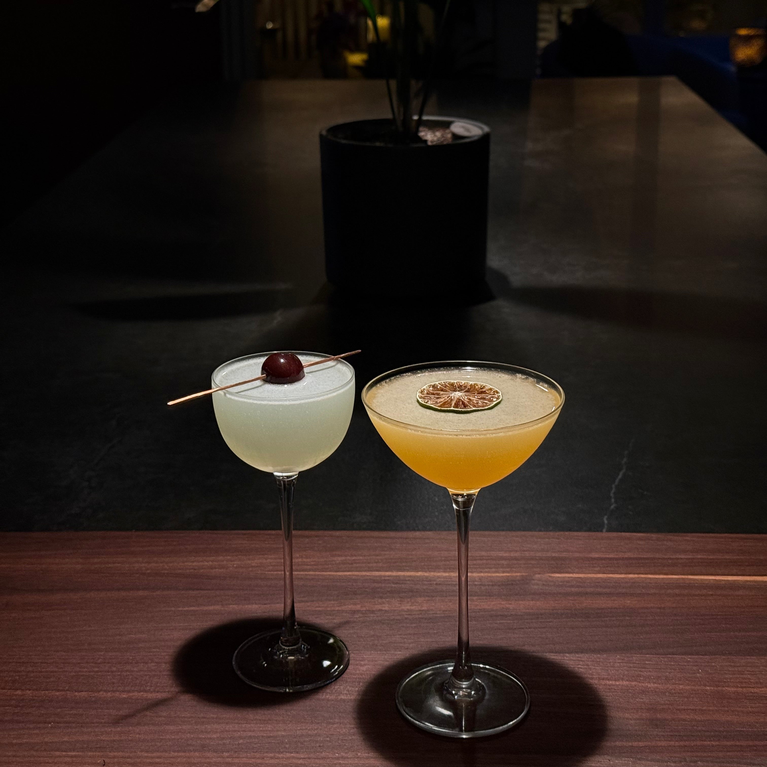 Two cocktails in stemmed glasses on a dark tabletop in a dimly lit bar or restaurant setting. One drink is pale creamy white with a cherry garnish, and the other is yellow with a spoonful of foam on top.