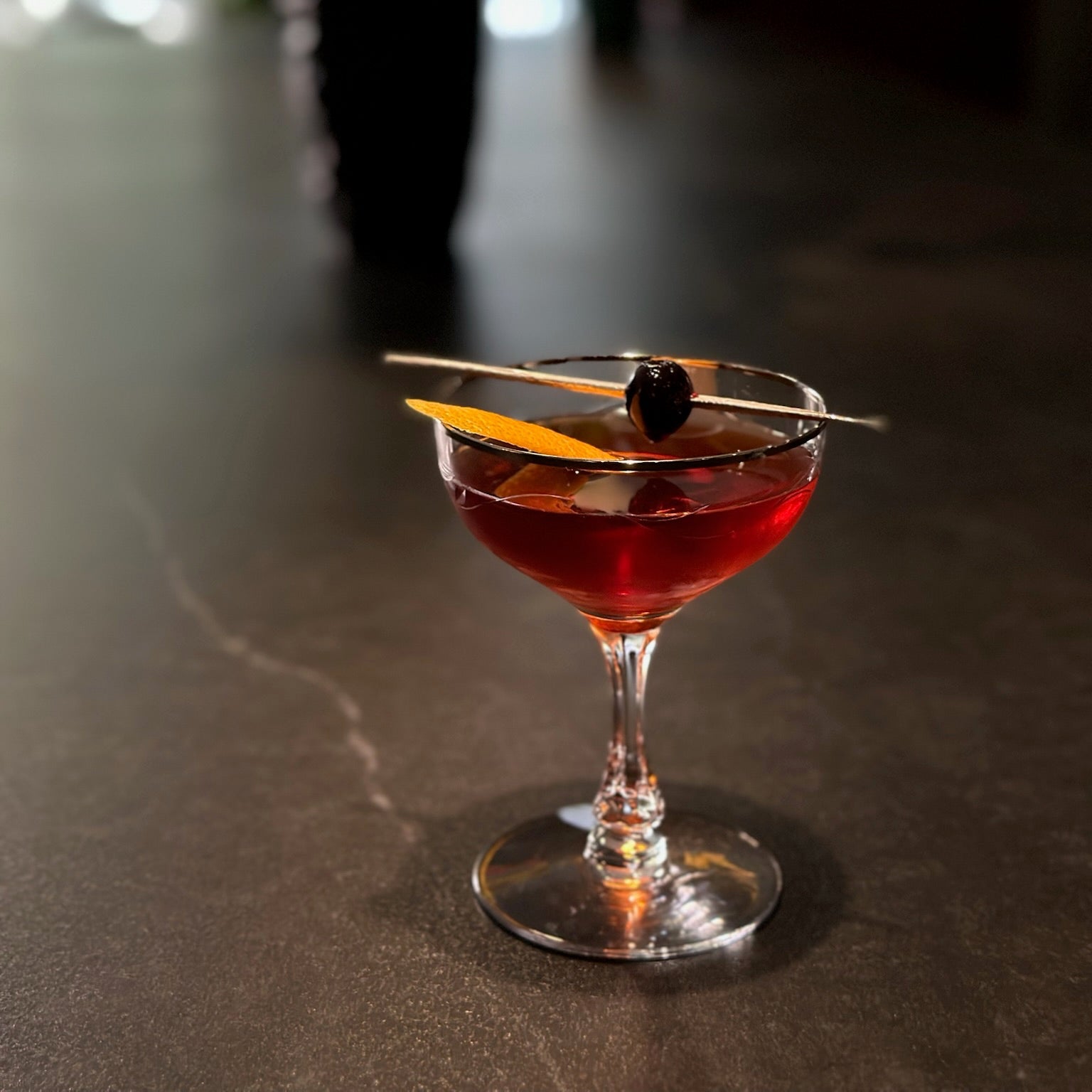 Red cocktail in a glass with garnishes on a dark surface, blurred flowers in the background