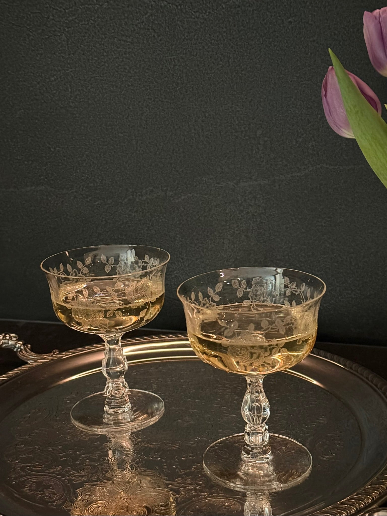 Two glass coupes with a golden liquid on a reflective surface with a dark background.