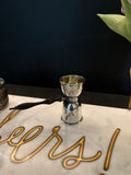 A polished silver metal double jigger or measuring pour with a black wood handle, set on a white marble tabletop with gold decorative script and a dark background.