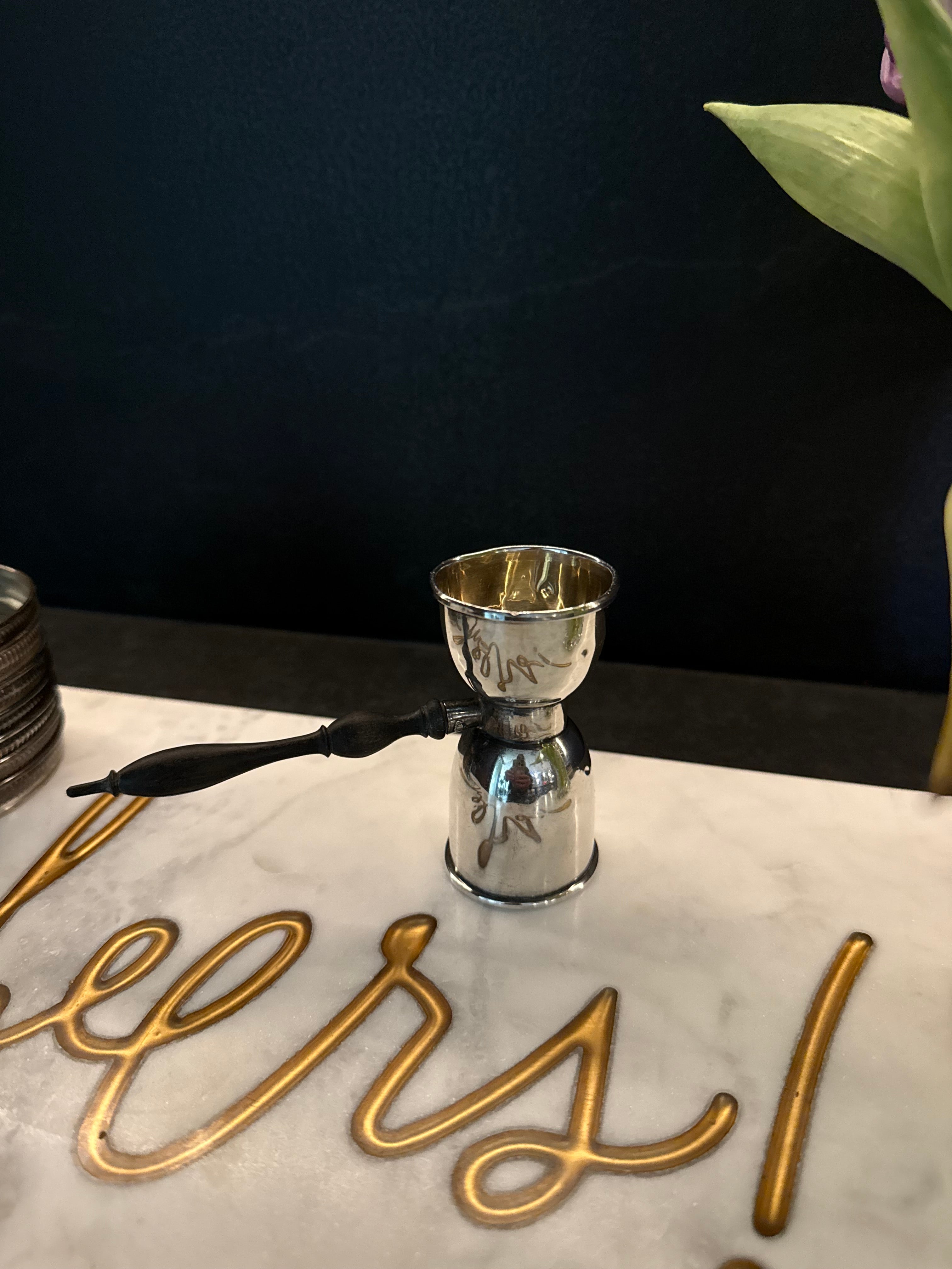 A polished silver metal double jigger or measuring pour with a black wood handle, set on a white marble tabletop with gold decorative script and a dark background.