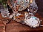 A silver bar tray arranged on a wooden table with a jigger, small white square dish, copper-toned bar tools in a glass holder, a strainer spoon, and a clear glass decanter in the background.