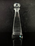 A clear glass decanter with a tapered body, horizontal ribbed detailing, and a spherical stopper, set against a dark textured background.