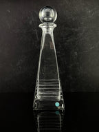 A clear glass decanter with a tapered body, horizontal ribbed detailing, and a spherical stopper, set against a dark textured background.