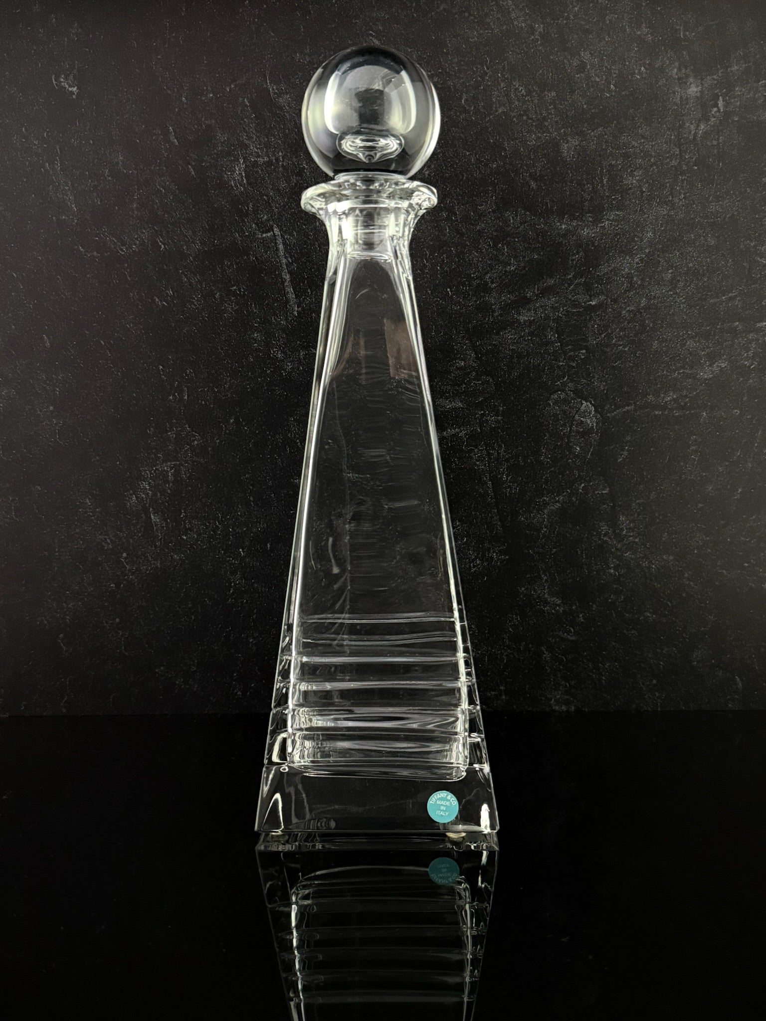 A clear glass decanter with a tapered body, horizontal ribbed detailing, and a spherical stopper, set against a dark textured background.