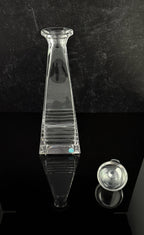 A clear glass decanter with a flared rim and ribbed body, shown on a glossy black surface against a dark textured background, with a small detached glass stopper visible to the right.