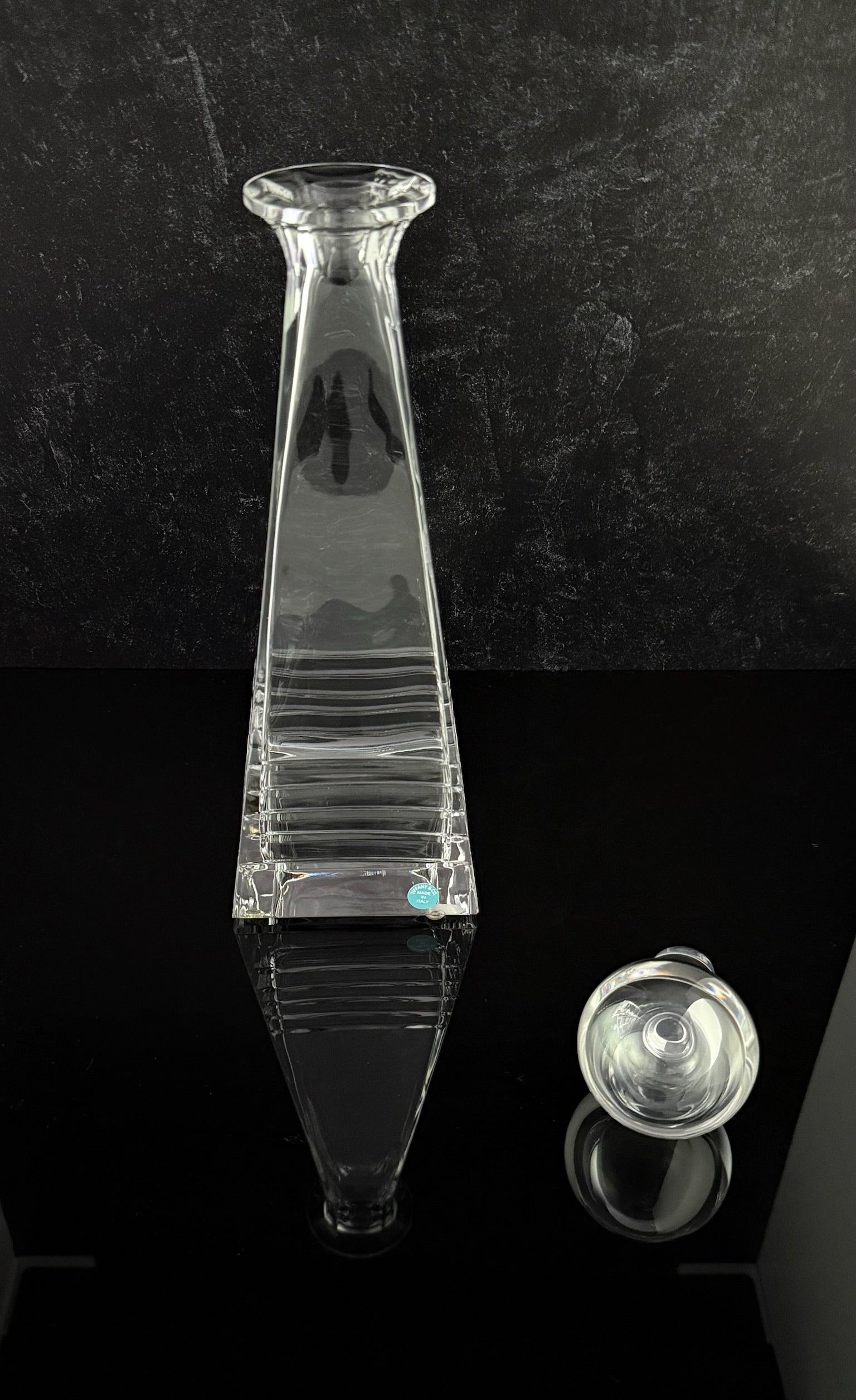 A clear glass decanter with a flared rim and ribbed body, shown on a glossy black surface against a dark textured background, with a small detached glass stopper visible to the right.