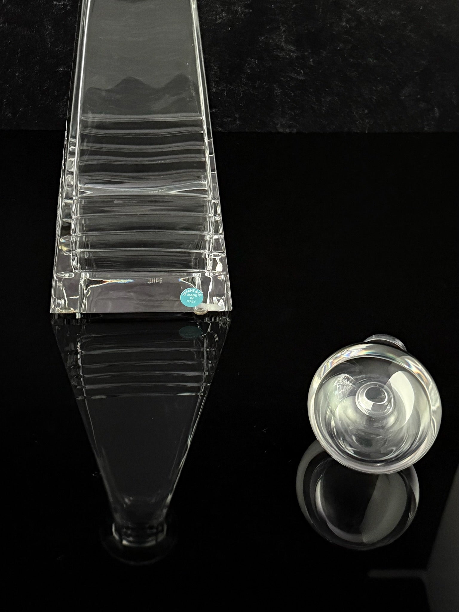 A closeup photo of crystal decanter base with horizontal ridges, shown alongside a matching round clear stopper on a glossy black reflective surface.