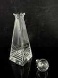 A clear glass decanter with a geometric, faceted design and horizontal ridges near the base, shown next to its matching glass stopper on a reflective black surface against a dark textured background.