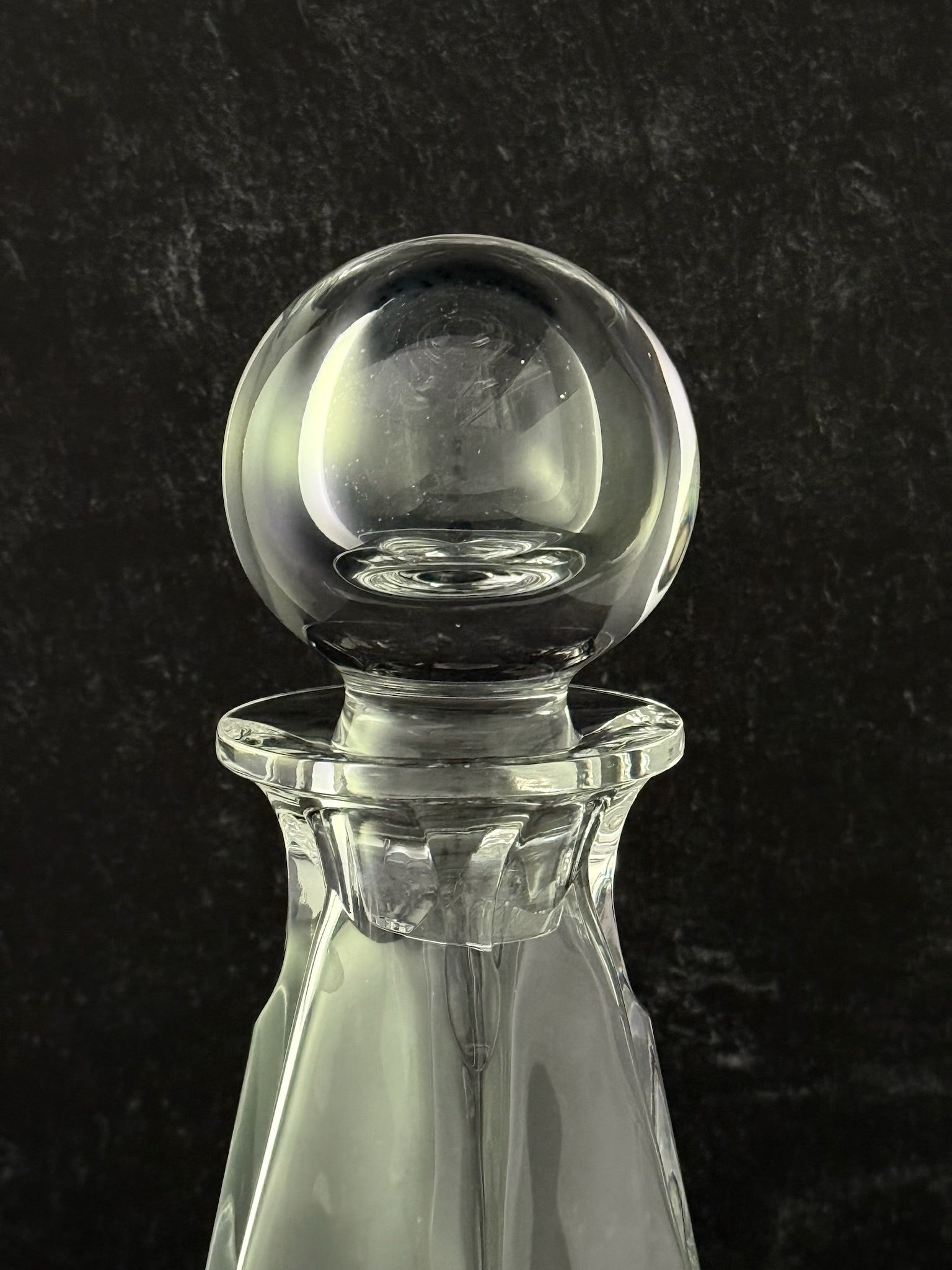 A clear glass decanter with a spherical glass stopper, shown against a dark textured background.