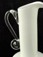 A close-up of a white glass vessel with a clear curved handle and a small bubble detail, set against a black background.