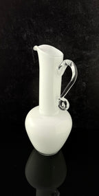 A white glass pitcher with a curved, clear glass handle, set against a dark black background with a reflective surface below.