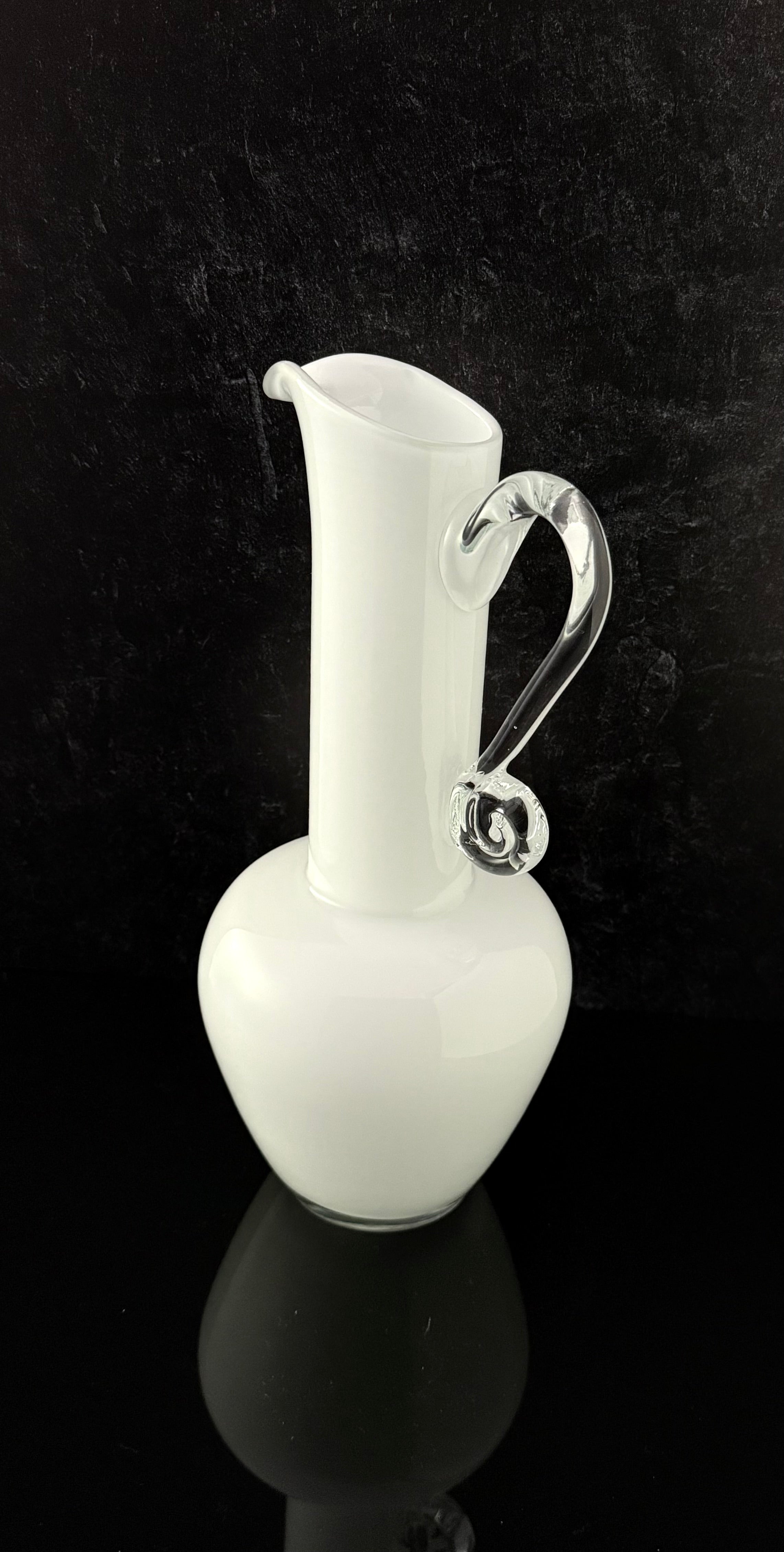 A white glass pitcher with a curved, clear glass handle, set against a dark black background with a reflective surface below.