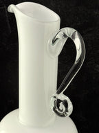 A white glass pitcher with a curved, clear glass handle, shown against a dark black background.