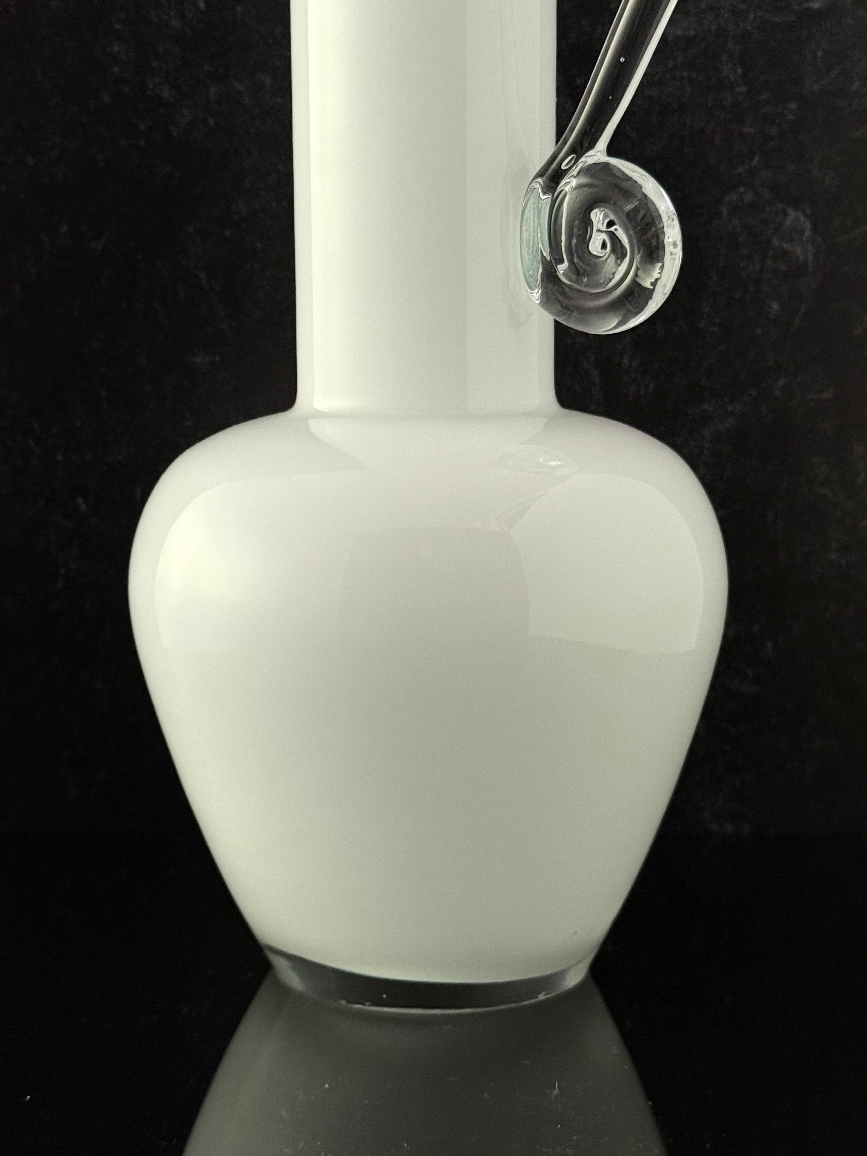 A white glass bottle or vase with a bulbous rounded body, a narrow neck, and a clear glass curved attachment near the top, photographed against a black background.