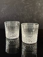 Two clear cut-crystal tumbler glasses with a textured patterned lower half, standing on a reflective black surface against a dark gray textured background.