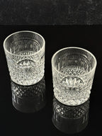 Two clear cut-crystal tumbler glasses with a floral pattern, displayed on a reflective black surface against a dark background.