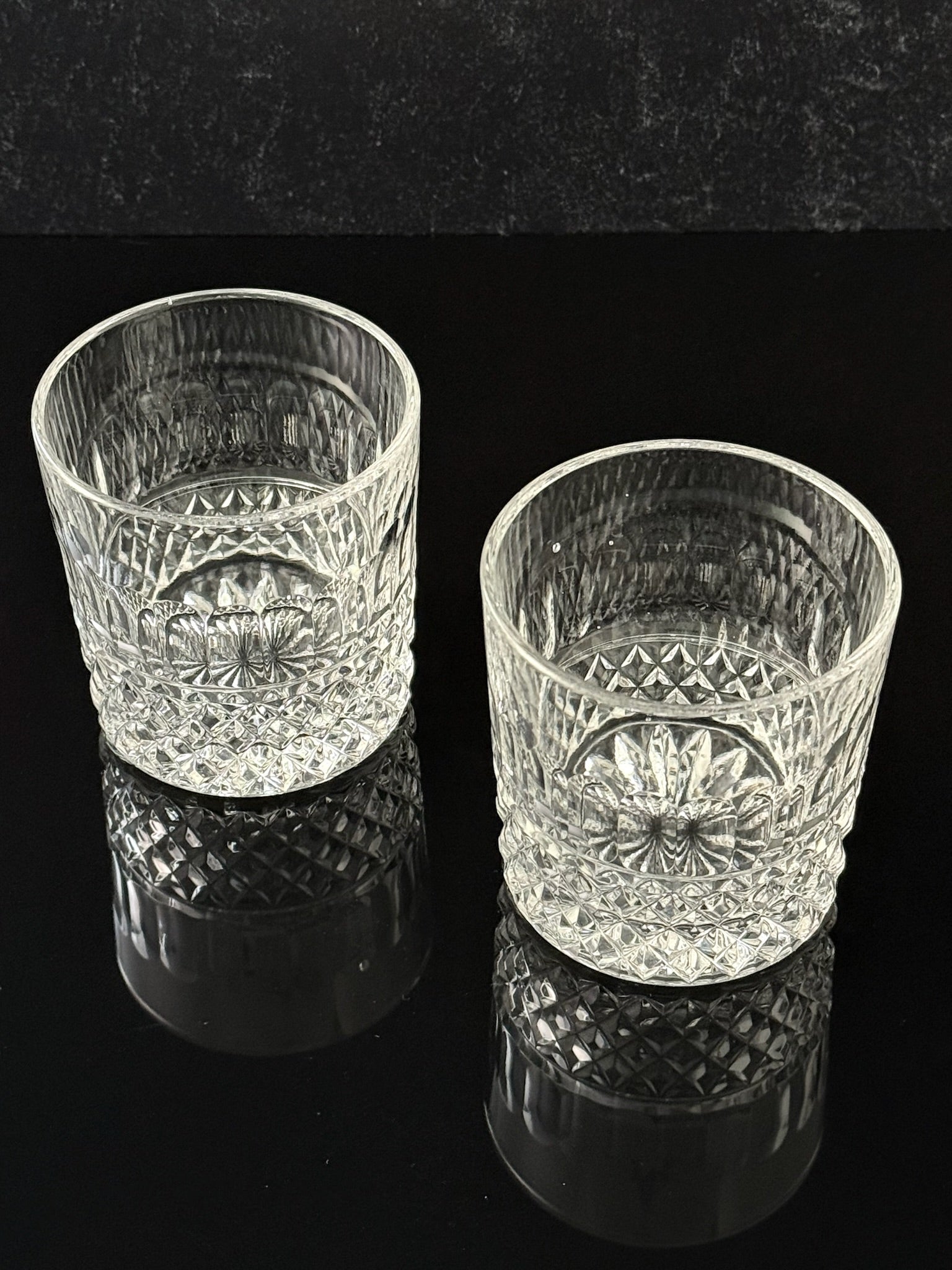 Two clear cut-crystal tumbler glasses with a floral pattern, displayed on a reflective black surface against a dark background.