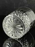 A clear cut-crystal tumbler glass with a faceted starburst pattern on the base, photographed on a dark reflective surface.