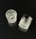 Two clear cut-crystal whiskey glasses with faceted patterns, displayed on a reflective black surface against a black background.