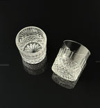 Two clear cut-crystal whiskey glasses with faceted patterns, displayed on a reflective black surface against a black background.