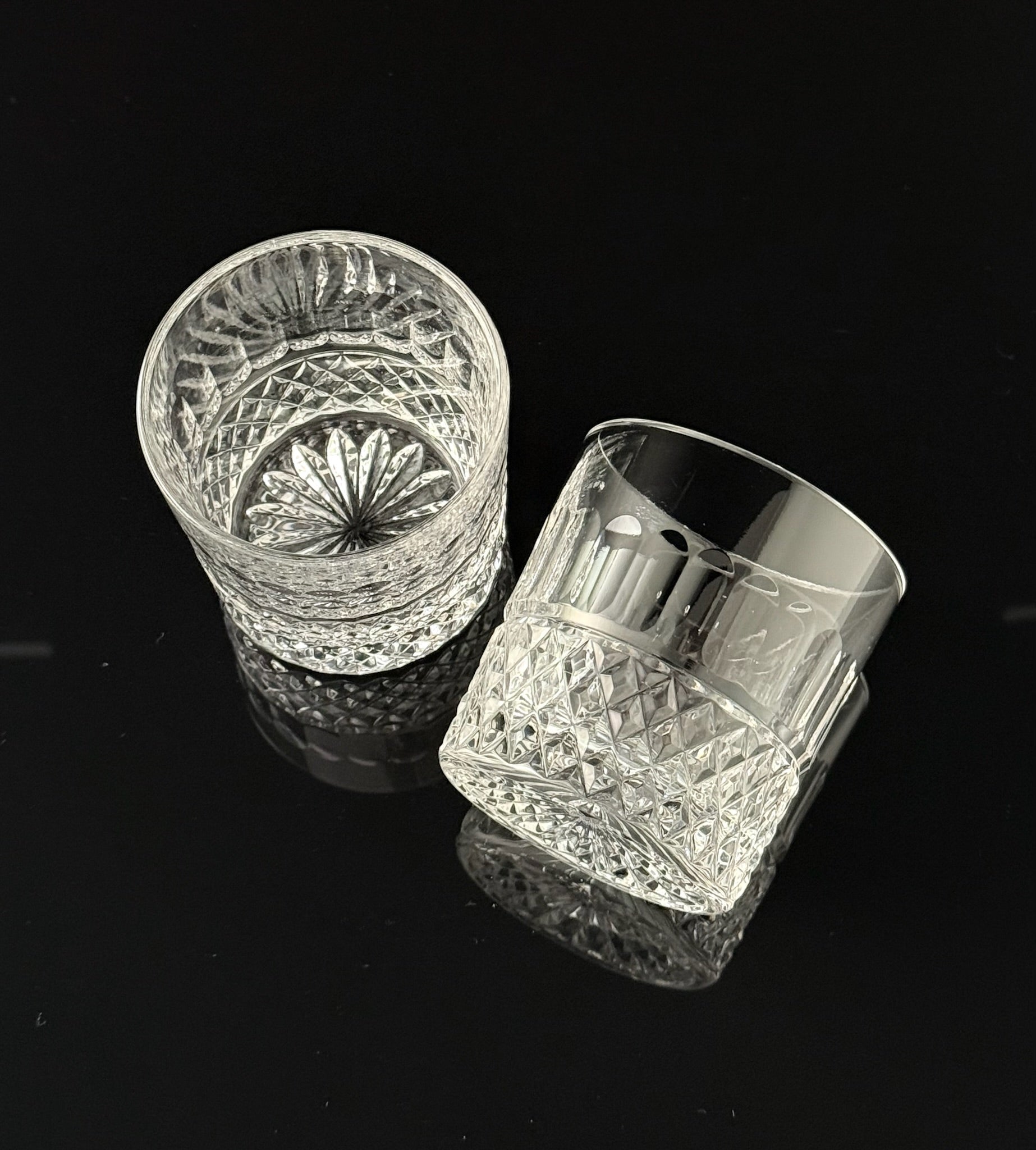 Two clear cut-crystal tumbler glasses with faceted geometric patterns, displayed on a reflective black surface.