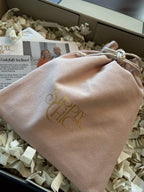 A blush pink velvet drawstring pouch with gold foil 'Sipply Chic' branding, nestled in crinkle paper inside a gift box.