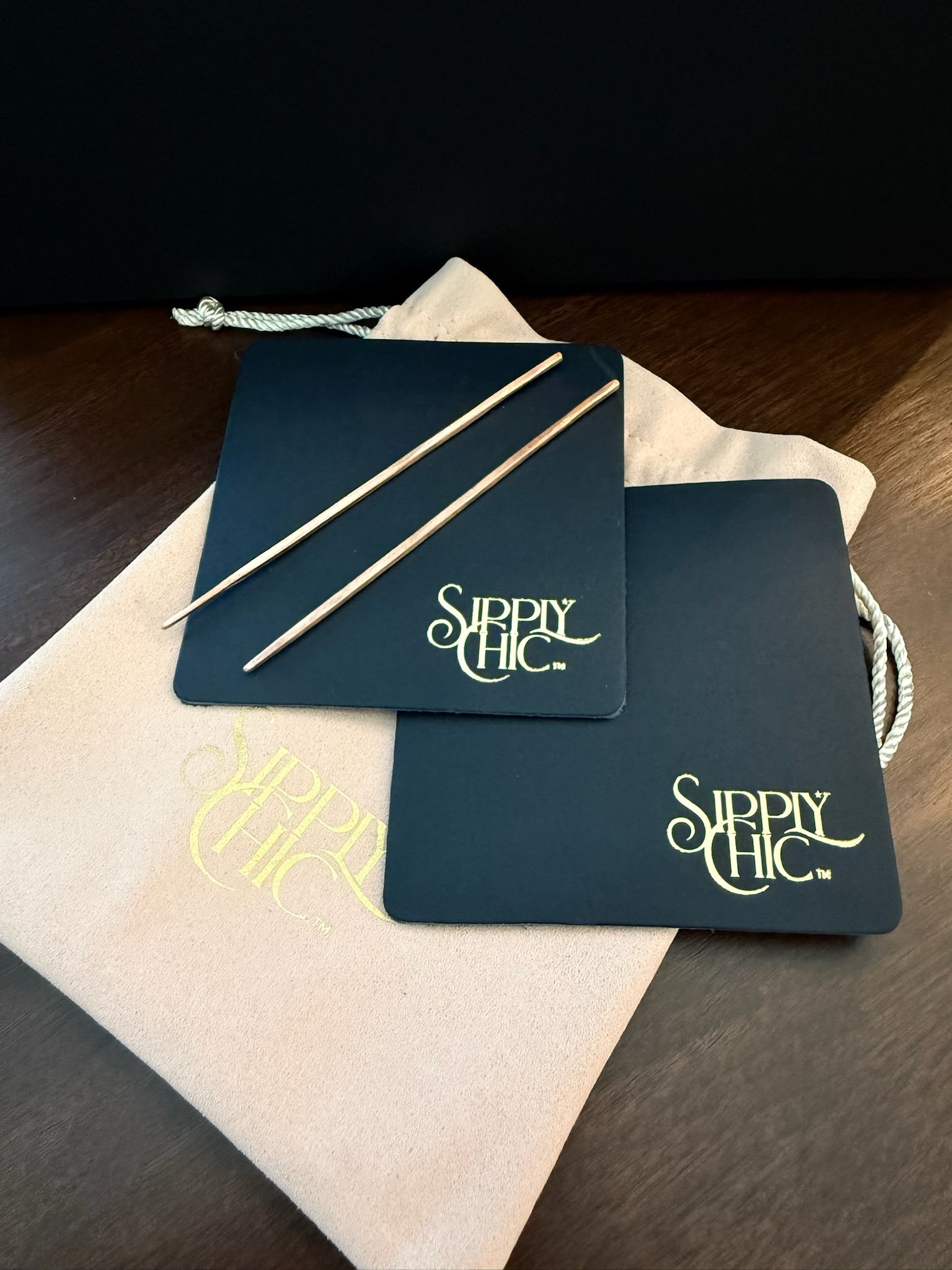 Two navy rectangular coasters with gold 'Sipply Chic' logos and two thin copper cocktail picks, arranged on top of a rose colored drawstring pouch on a dark wooden surface.