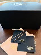 A black Sipply Chic gift box sits on a wooden table with a rose colored pouch, two navy leather coasters, and two copper cocktail picks in front. The box lid and coasters features the Sipply Chic logo in gold.