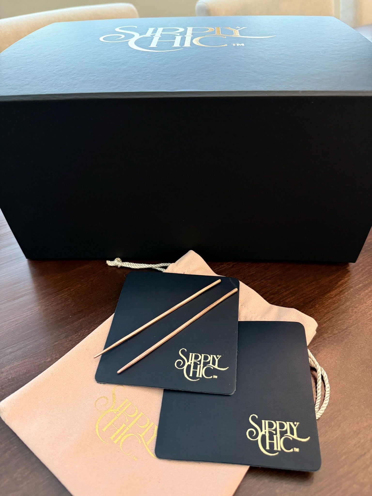 A black Sipply Chic gift box sits on a wooden table with a rose colored pouch, two navy leather coasters, and two copper cocktail picks in front. The box lid and coasters features the Sipply Chic logo in gold.