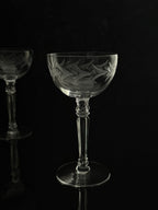 A clear cut-glass coupe glass with etched floral and leaf patterns, standing on a reflective black surface against a dark background. A second matching glass is partially visible in the out-of-focus background on the left.