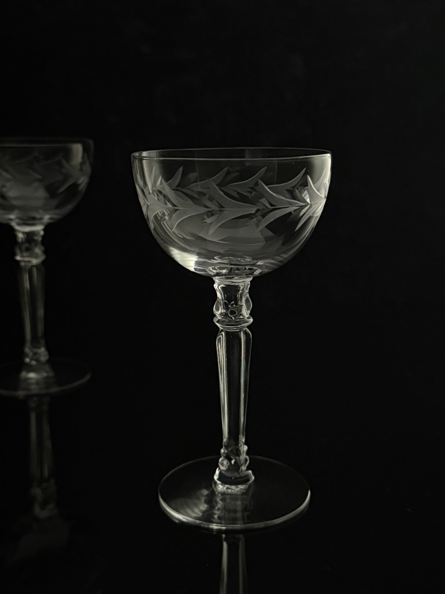 A clear cut-glass coupe glass with etched floral and leaf patterns, standing on a reflective black surface against a dark background. A second matching glass is partially visible in the out-of-focus background on the left.