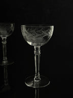 A clear cut-glass coupe-style wine glass with etched floral and leaf patterns, standing on a reflective black surface against a black background. A second matching glass is partially visible in the out-of-focus background on the left.