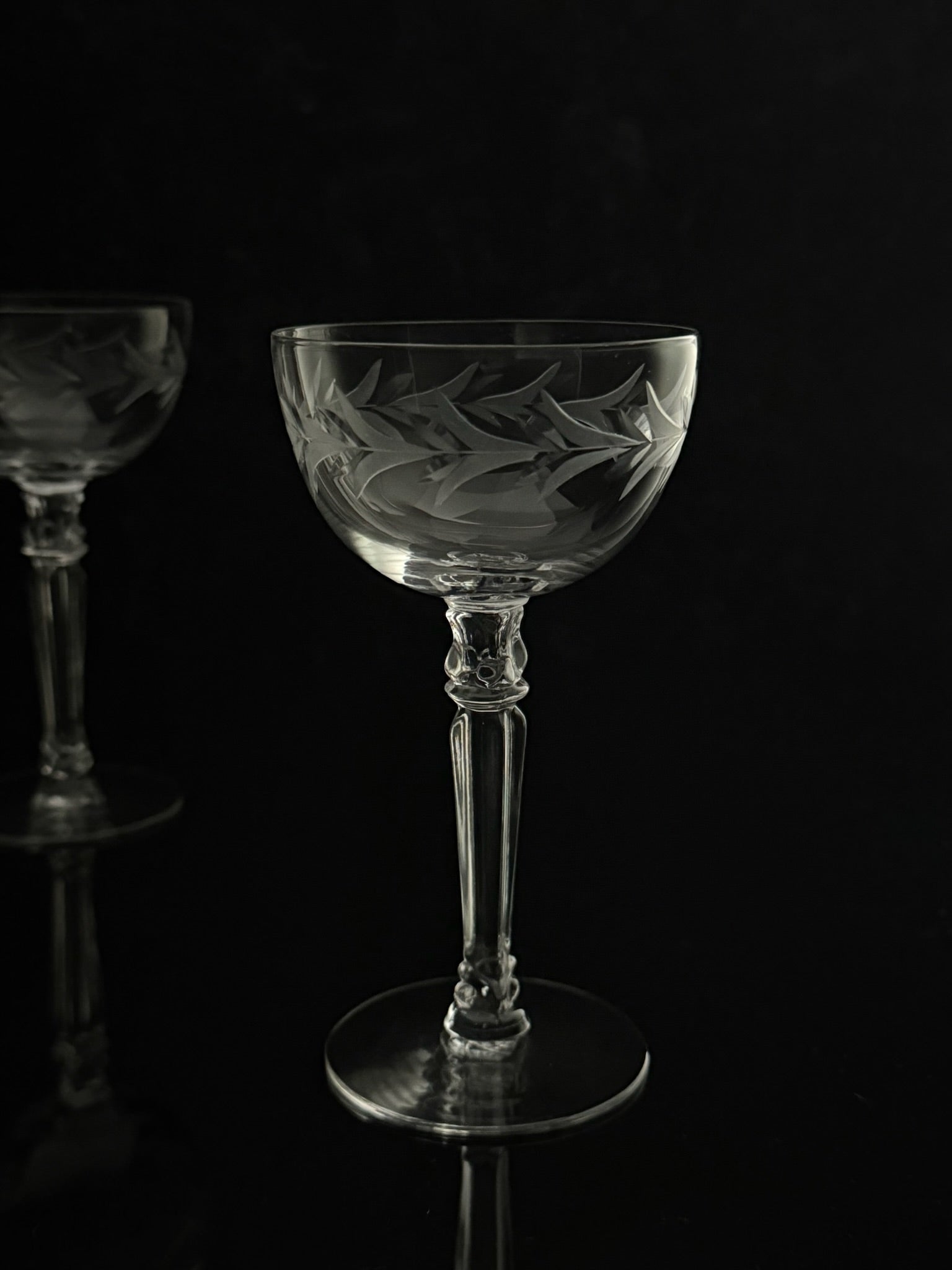 A clear cut-glass coupe-style wine glass with etched floral and leaf patterns, standing on a reflective black surface against a black background. A second matching glass is partially visible in the out-of-focus background on the left.