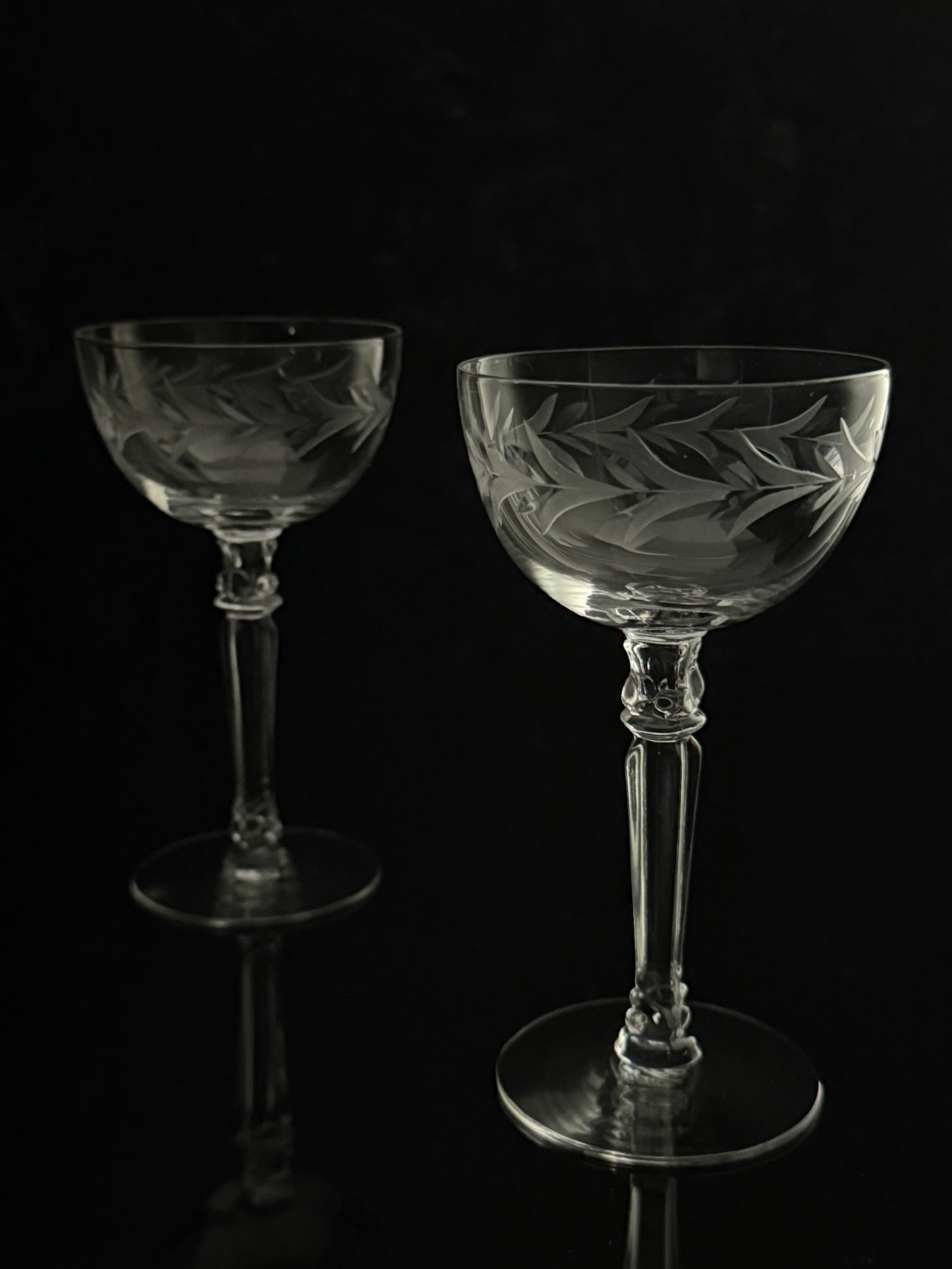 Two clear cut-glass coupe glasses with etched floral patterns, standing on a reflective black surface against a black background.