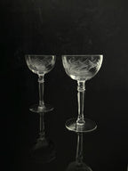 Two clear cut-glass coupe glasses with etched floral patterns, standing on a reflective black surface against a dark background.