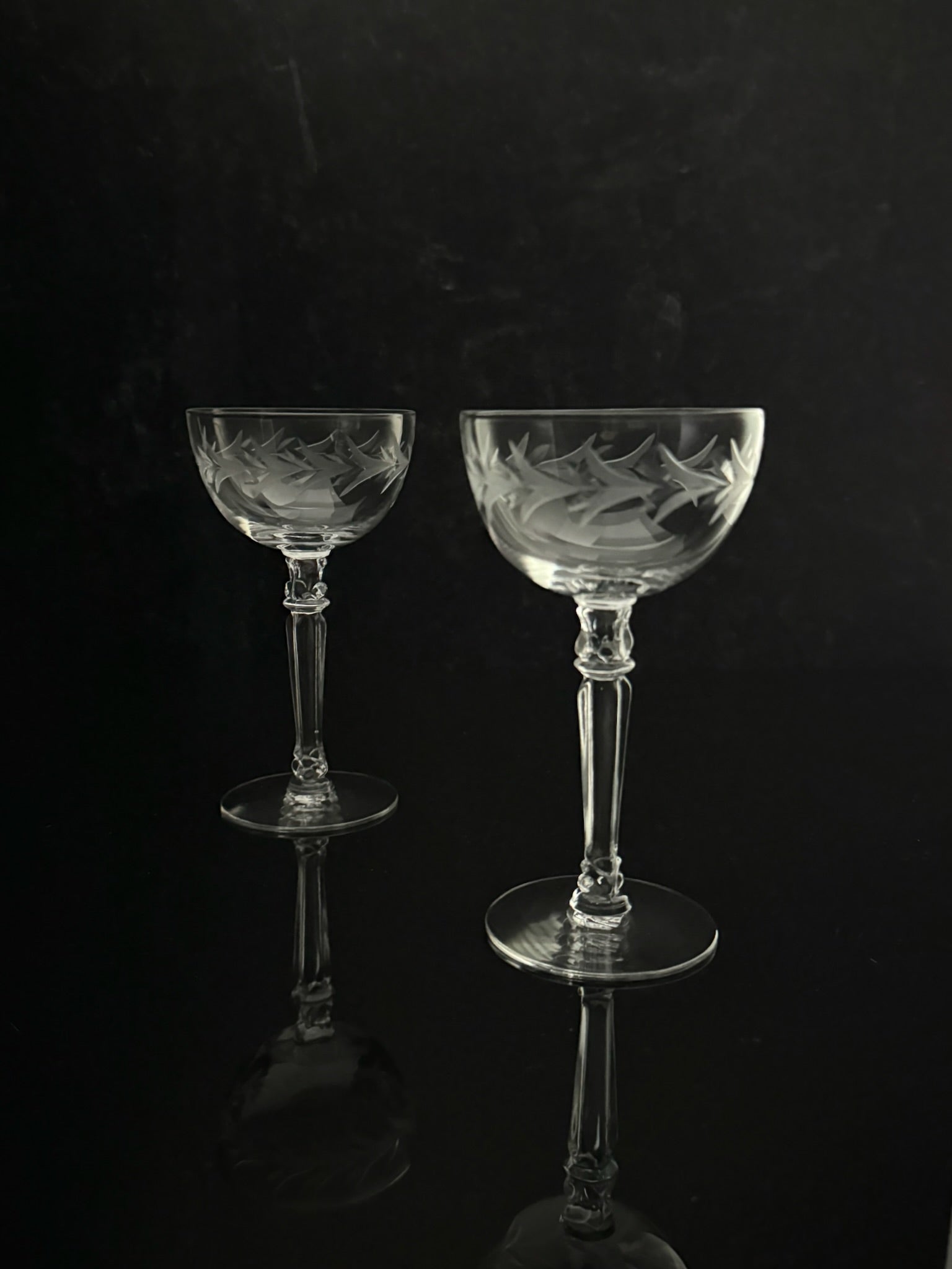 Two clear cut-glass coupe glasses with etched floral patterns, standing on a reflective black surface against a dark background.
