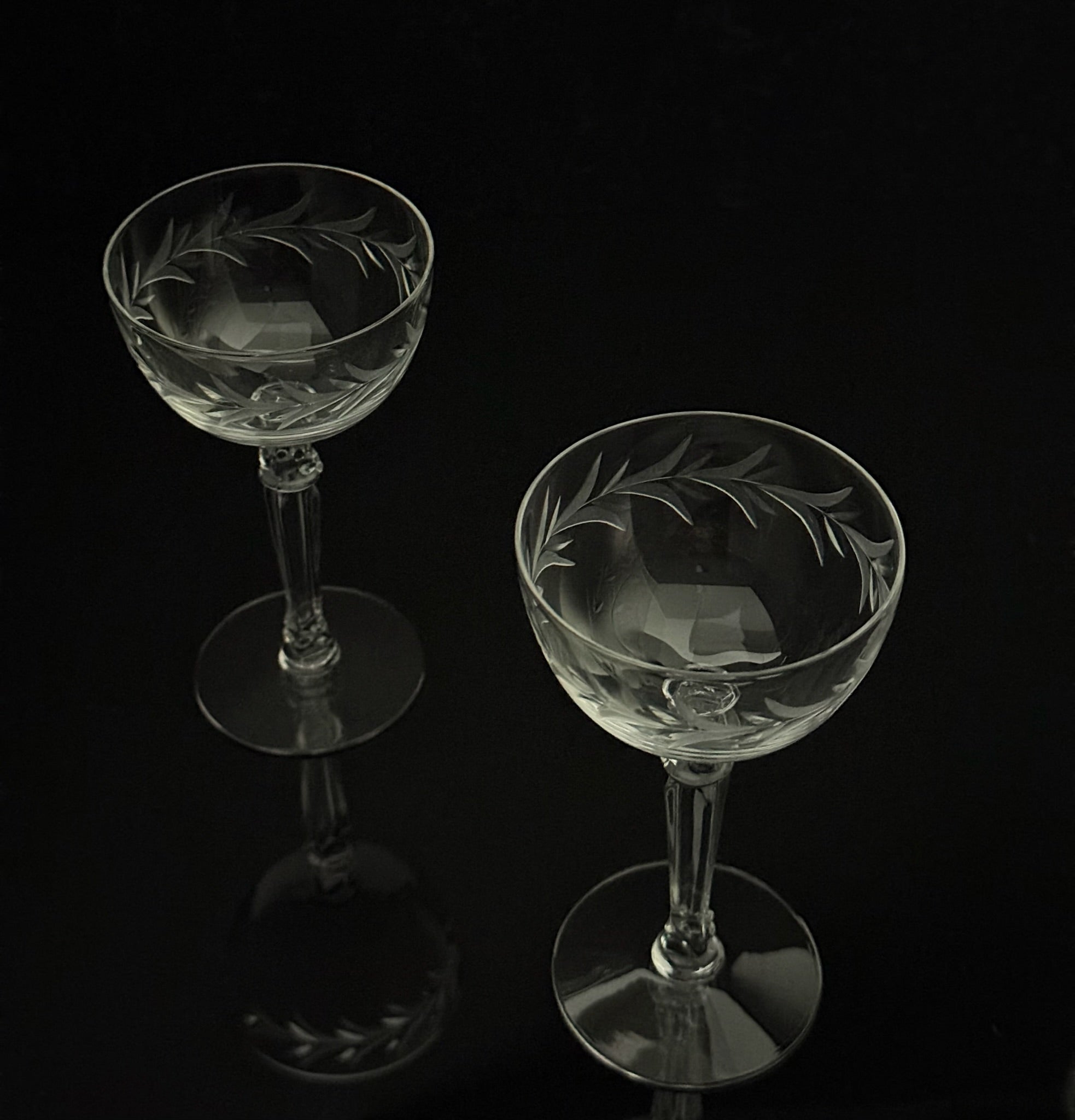 Two clear crystal coupe glasses with etched leaf patterns, displayed on a reflective black surface against a black background.