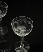 A clear cut-glass coupe glass with etched leaf patterns, shown on a reflective black background with another glass partially visible at the upper left.