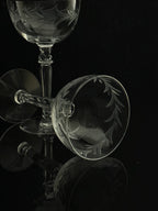 Clear etched glass wine glasses with leaf motifs, arranged on a reflective black surface against a dark background.
