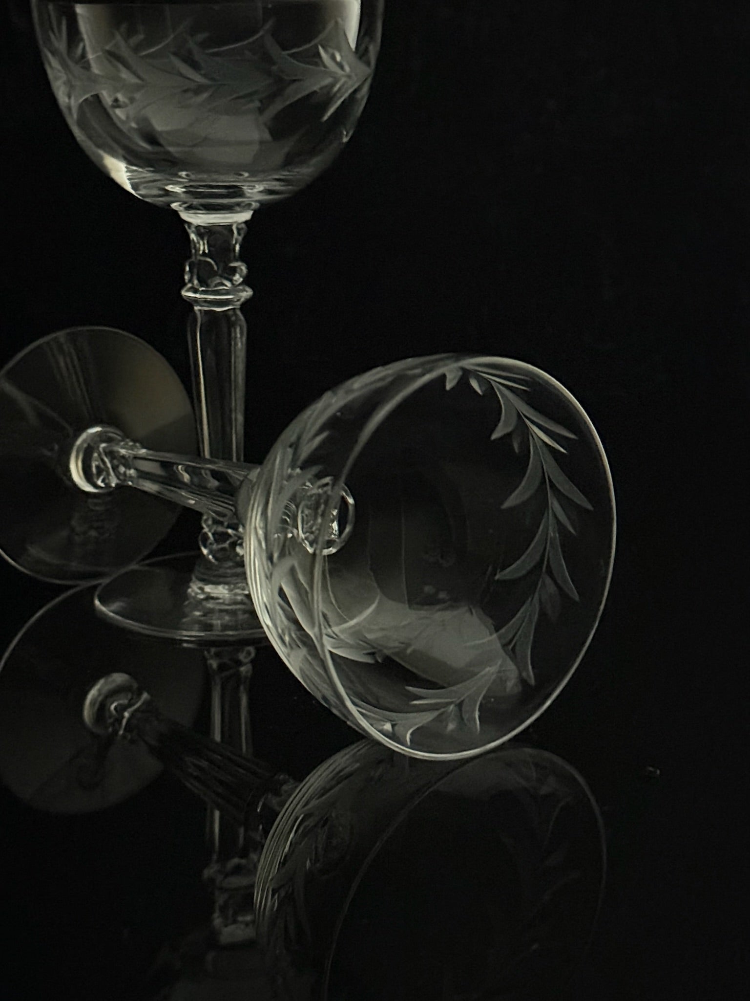 Clear etched glass wine glasses with leaf motifs, arranged on a reflective black surface against a dark background.