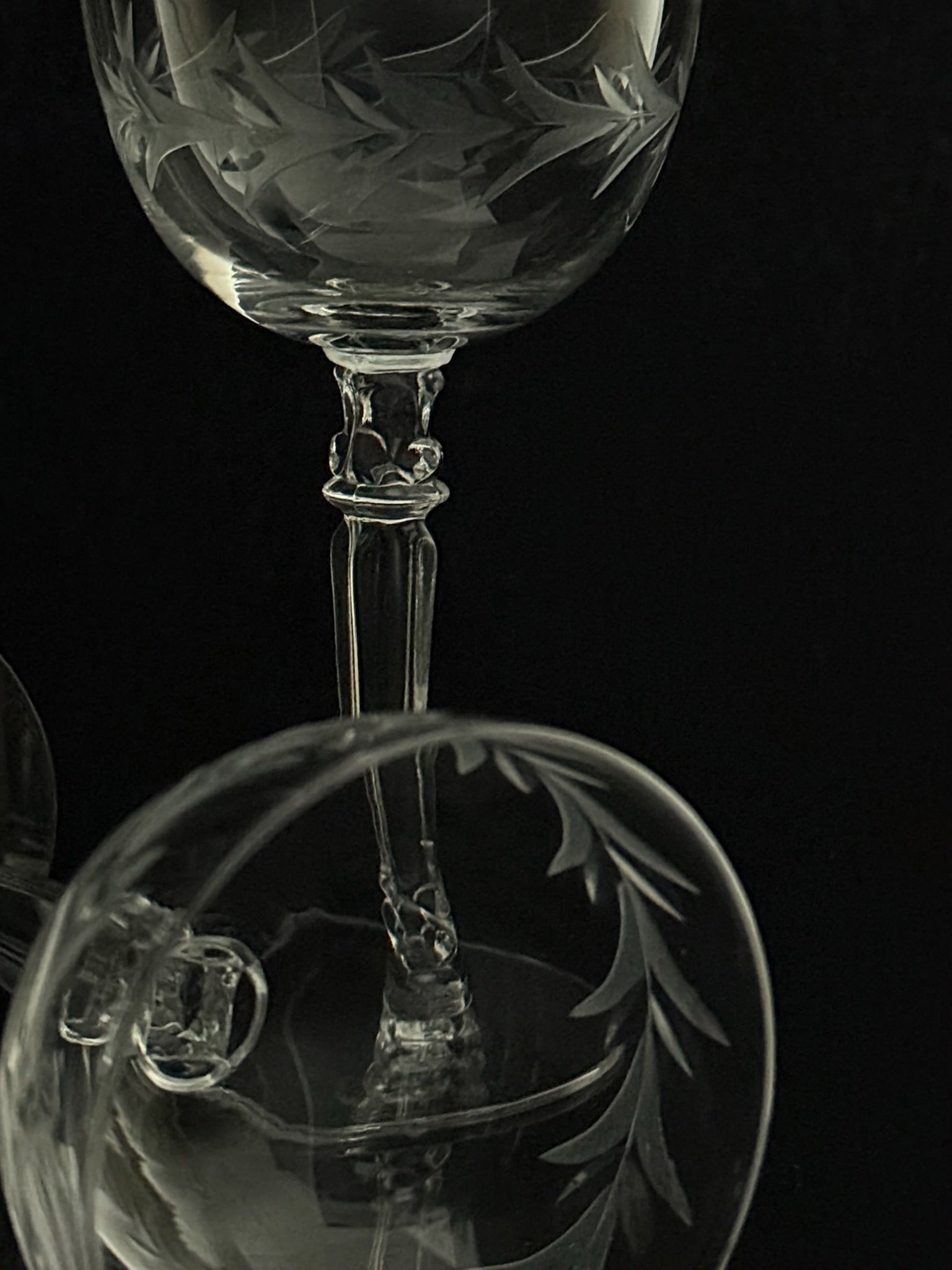 Close-up studio shot of a clear etched glass wine glass against a black background, showing the bowl and stem with decorative leaf-like engraving.